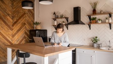 Canva - Busy female freelancer with laptop taking notes in kitchen