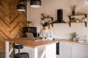 Canva - Busy female freelancer with laptop taking notes in kitchen