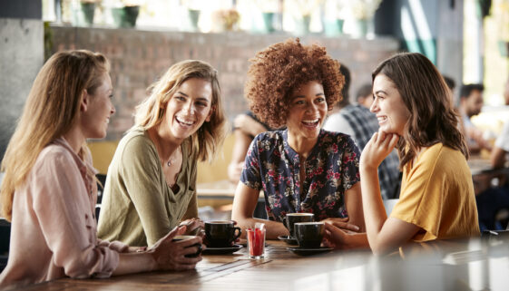 Four Young Female Friends Meeting Sit At Table In Coffee Shop And Talk