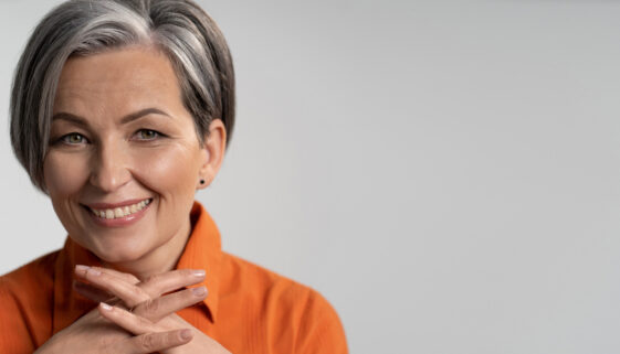 Mature woman broadly smiling with fingers crossed. Happy female model in orange shirt. Close up portrait. Maturity concept. Horizontal template with empty place at right side