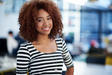 Going places. Beautiful young woman smiling at the camera with open office space behind her.