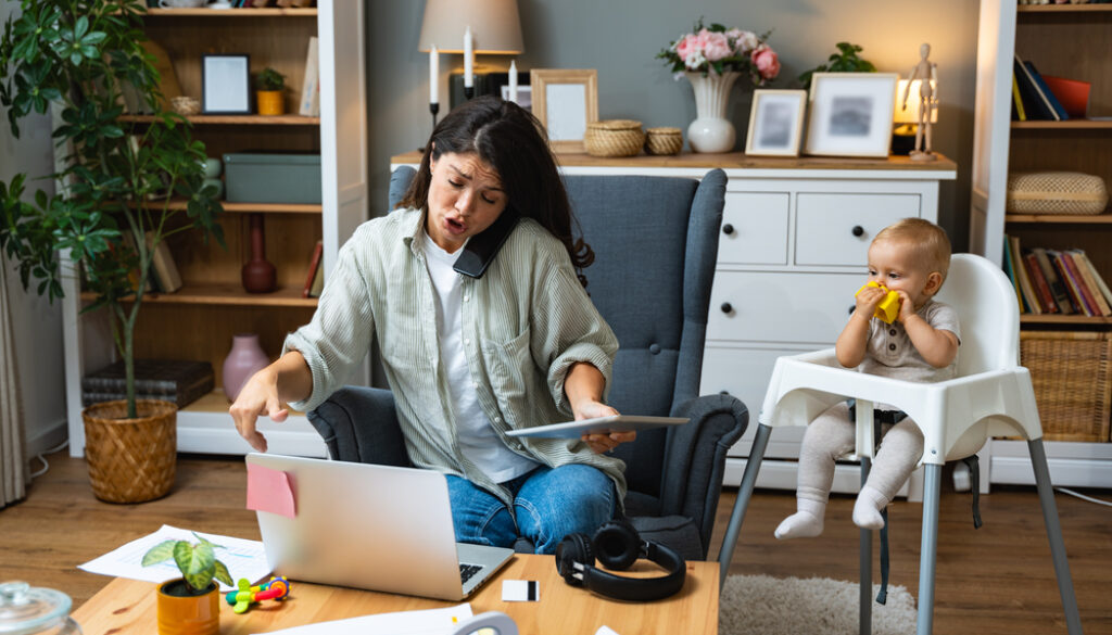 Mother working on laptop and talking on computer webcam video call communication with business partners while baby playing. Woman work from home and caring for kids.