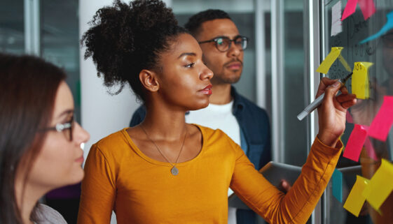 Young creative businesswoman standing with her colleagues writing new ideas on sticky notes over glass wall