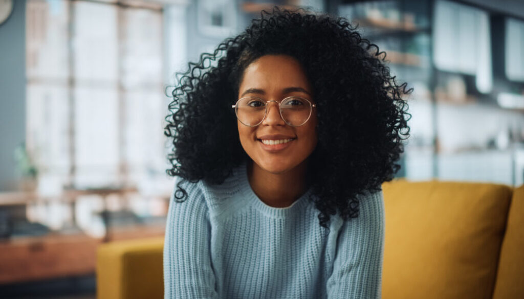 Portrait of a Beautiful Authentic Latina Female with Afro Hair Wearing Light Blue Jumper and Glasses. She Looks to the Camera and Smiling Charmingly. Successful Woman Resting in Bright Living Room.