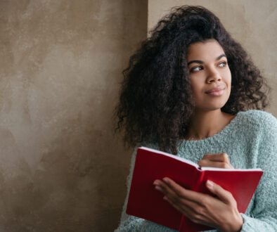 Beautiful African American woman holding red book, looking at window and smiling. University student studying, learning language, sitting at library. Portrait of young pensive writer taking notes