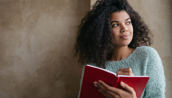 Beautiful African American woman holding red book, looking at window and smiling. University student studying, learning language, sitting at library. Portrait of young pensive writer taking notes